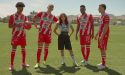 Eva Longoria stands confidently on a soccer field with four Club Necaxa players wearing their red and white team uniforms. The players pose with a soccer ball as mountains and training facilities are visible in the background. This promotional image is for the upcoming docuseries “Necaxa.”