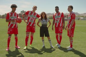 Eva Longoria stands confidently on a soccer field with four Club Necaxa players wearing their red and white team uniforms. The players pose with a soccer ball as mountains and training facilities are visible in the background. This promotional image is for the upcoming docuseries “Necaxa.”