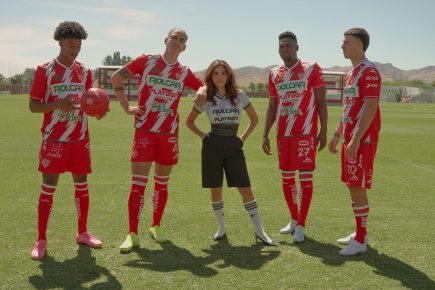 Eva Longoria stands confidently on a soccer field with four Club Necaxa players wearing their red and white team uniforms. The players pose with a soccer ball as mountains and training facilities are visible in the background. This promotional image is for the upcoming docuseries “Necaxa.”
