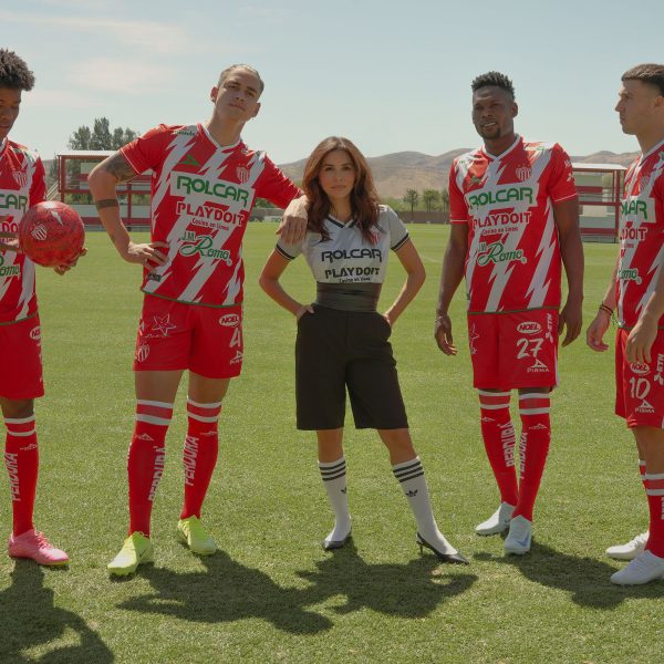 Eva Longoria stands confidently on a soccer field with four Club Necaxa players wearing their red and white team uniforms. The players pose with a soccer ball as mountains and training facilities are visible in the background. This promotional image is for the upcoming docuseries “Necaxa.”