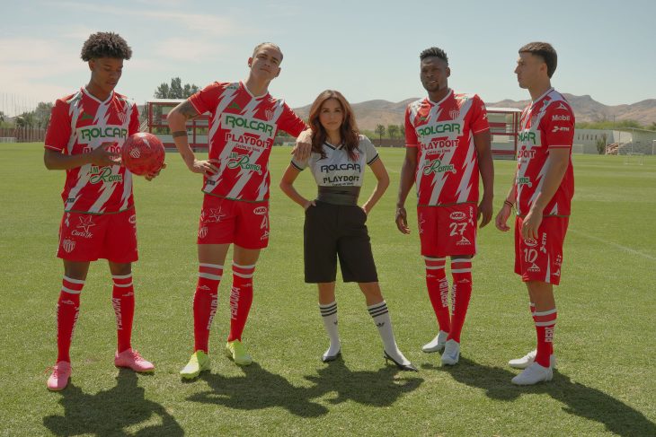Eva Longoria stands confidently on a soccer field with four Club Necaxa players wearing their red and white team uniforms. The players pose with a soccer ball as mountains and training facilities are visible in the background. This promotional image is for the upcoming docuseries “Necaxa.”