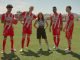 Eva Longoria stands confidently on a soccer field with four Club Necaxa players wearing their red and white team uniforms. The players pose with a soccer ball as mountains and training facilities are visible in the background. This promotional image is for the upcoming docuseries “Necaxa.”