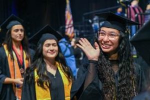 Hispanic female graduates in caps and gowns smile and wave during a graduation ceremony. The Tu Camino Scholarship supports Hispanic adults pursuing career growth through education. Credit: Lone Star College System.