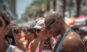 A crowd of people at an outdoor event in bright sunlight, their skin glistening with sweat. A man in the foreground wearing sunglasses and a sleeveless shirt holds a clear plastic cup and adjusts his glasses, while others around him shield their faces and drink cold beverages. Palm trees and colorful vendor tents are visible in the blurred background.