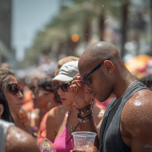 A crowd of people at an outdoor event in bright sunlight, their skin glistening with sweat. A man in the foreground wearing sunglasses and a sleeveless shirt holds a clear plastic cup and adjusts his glasses, while others around him shield their faces and drink cold beverages. Palm trees and colorful vendor tents are visible in the blurred background.